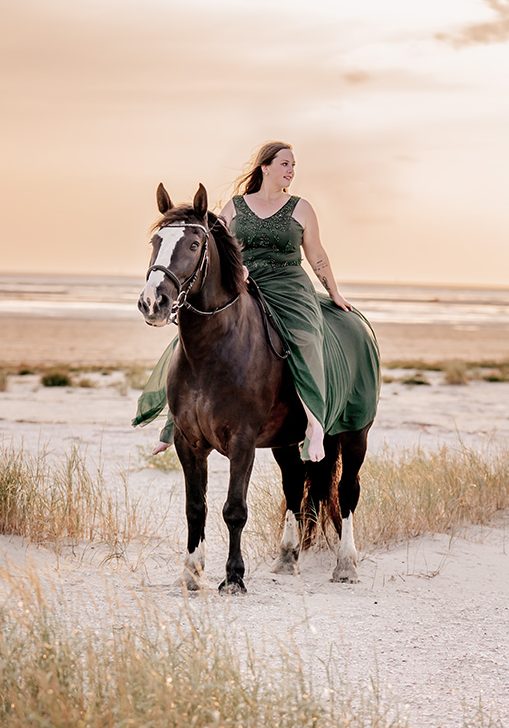 Frau in grünem Kleid reitet auf schwarzem Pferd am Strand bei Sonnenuntergang.