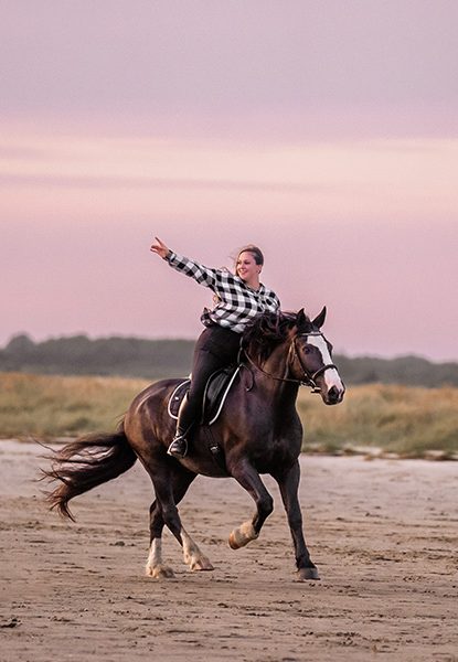Reiterin in kariertem Hemd auf einem Pferd bei Sonnenuntergang am Strand.