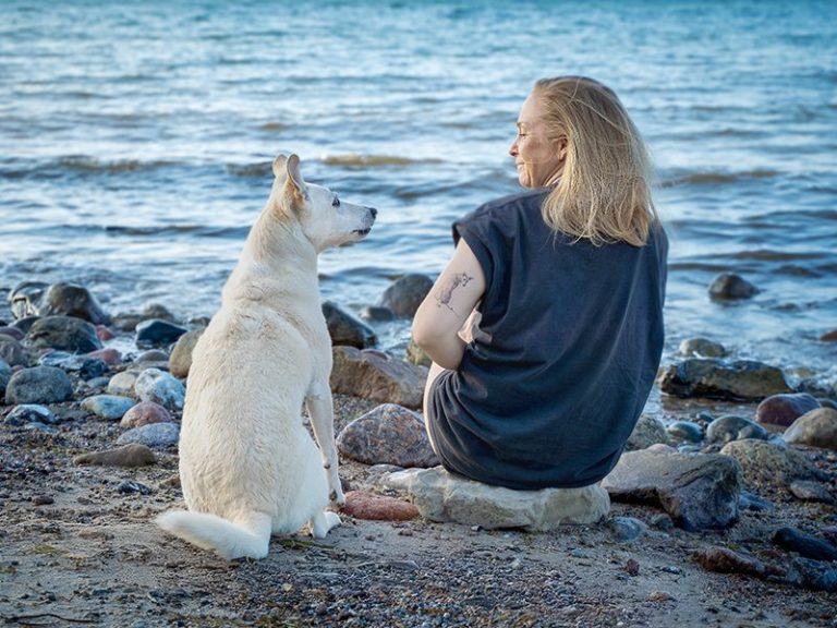 Eine Frau sitzt am Ufer und blickt auf ihren Hund.