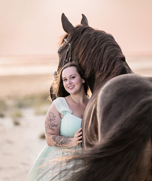 Frau in hellblauem Kleid umarmt ein schwarzes Pferd am Strand bei Sonnenuntergang.