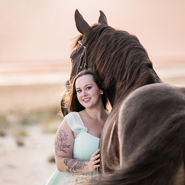 Frau mit Tätowierungen, die neben einem großen, braunen Pferd steht.