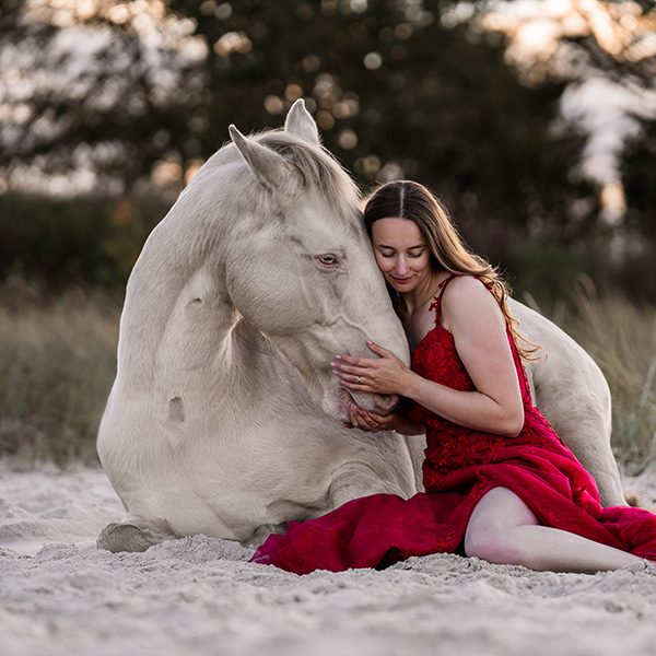 Frau in rotem Kleid umarmt ein weißes Pferd am Strand bei Sonnenuntergang.