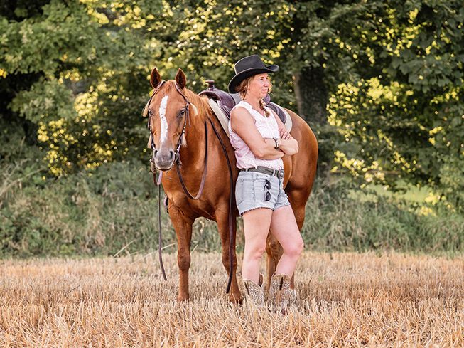 Frau in Shorts steht neben einem Pferd auf einem Stoppelfeld.