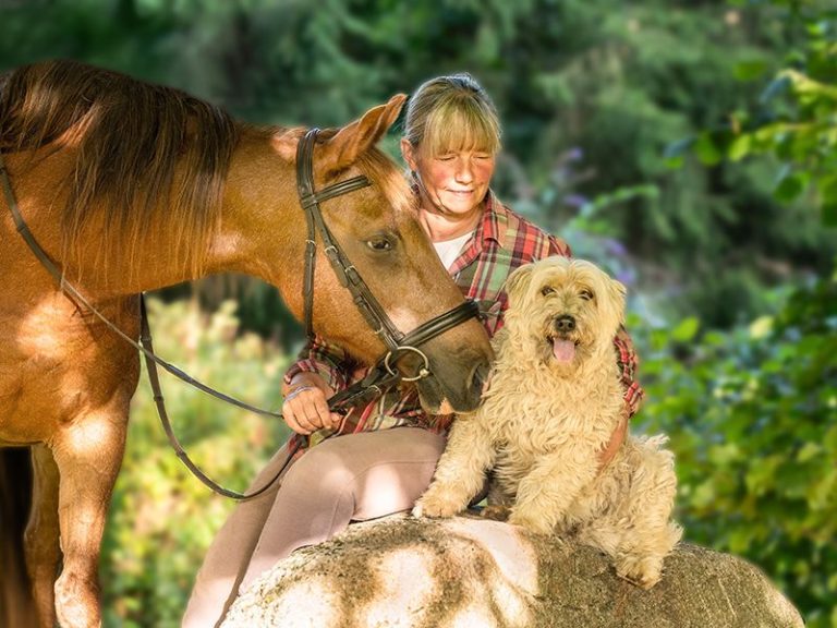 Eine Frau mit einem Hund sitzt auf einem Stein. Neben den beiden steht ein Pferd.