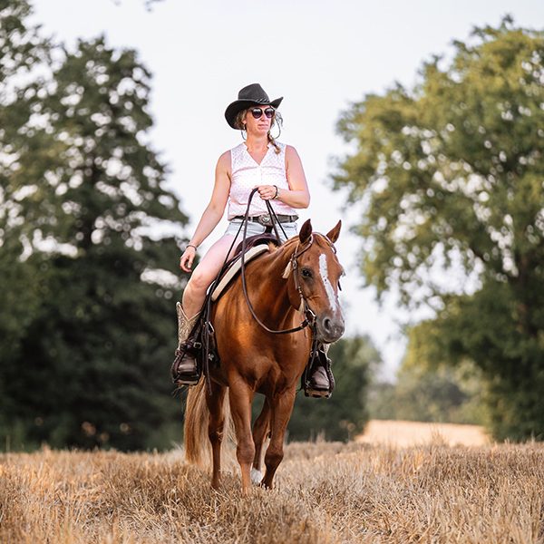 Reiterin mit Cowboyhut auf einem braunen Pferd (Quarter horse) in einem Feld.