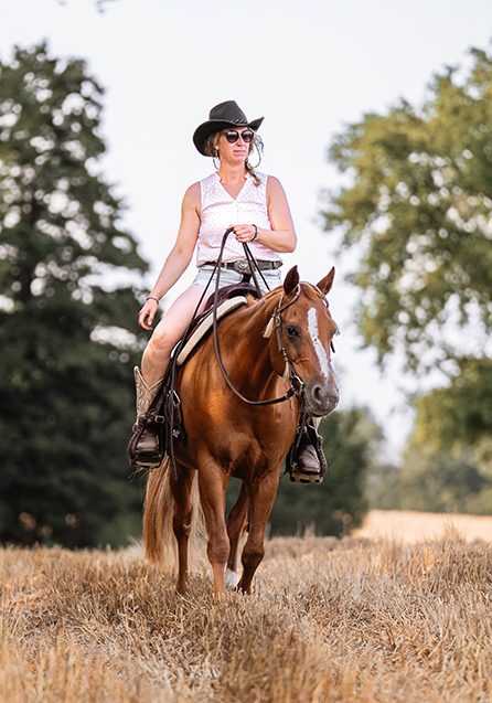 Westernreiterin mit Cowboy Hut auf braunem Pferd in einem Stoppelfeld.