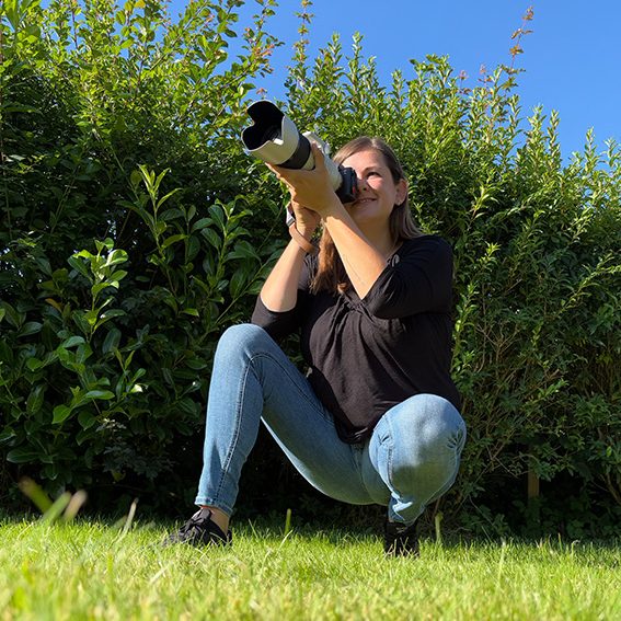 Die Fotografin kniet im Gras und fotografiert mit einer Kamera in Richtung des Betrachters.