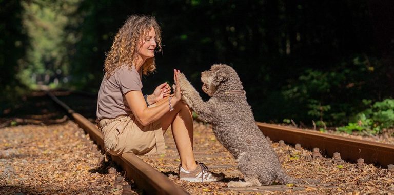Eine Frau sitzt bei sonnigem Wetter lächelnd auf den Gleisen, spielt mit einem Hund.