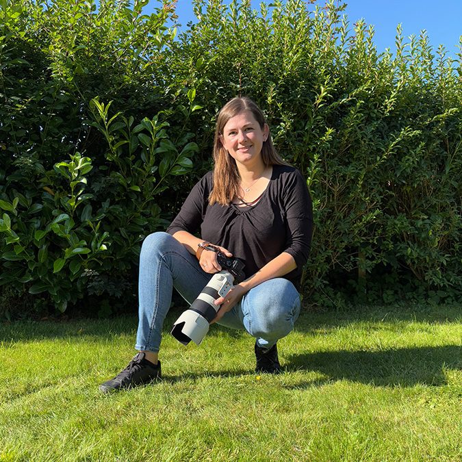 Frau hockt im Grünen, hält eine Kamera und trägt lässige Kleidung.